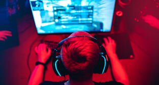 Imagem de um gamer jogando no computador. A tela do computador está destacada em azul e o resto da imagem tem um filtro vermelho. O gamer usa um fone de ouvido.